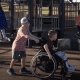 A young boy with CP rolls across a playground in his wheelchair while helped by another young boy pushing the handlebars.